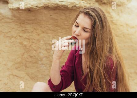 Beautiful blond girl in purple dress eating fresh strawberry on nature Stock Photo