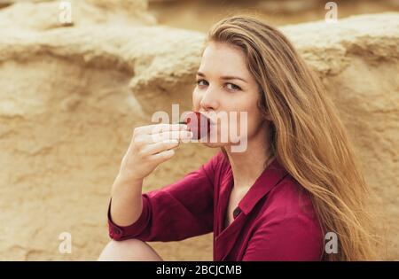 Beautiful blond smiling girl in purple dress eating fresh strawberry on nature Stock Photo