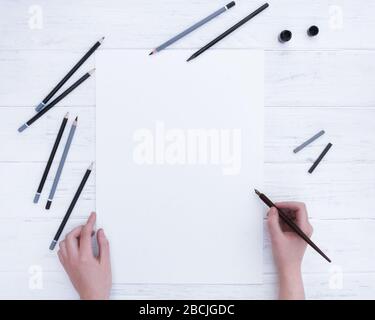 Mockup for calligraphy.  Hands holding the empty white paper sheet, and a calligraphy pen. A few pencils and black compressed charcoal.  View from ab Stock Photo