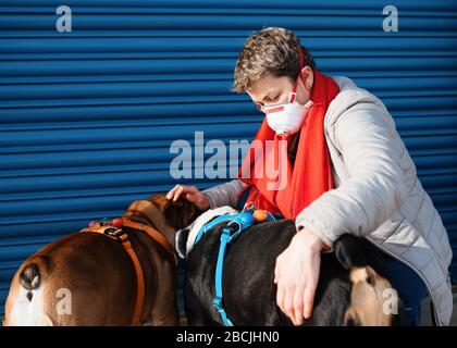 Portrait of woman wearing a white and red face mask, grey jacket and red scarf, stroking and hugging dogs against a blue background Stock Photo