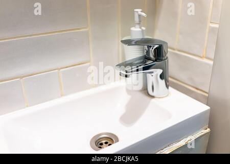 Modern bathroom stainless steel metal faucet in small tiny studio apartment sink closeup on tiles tiled wall of room and soap dispenser Stock Photo