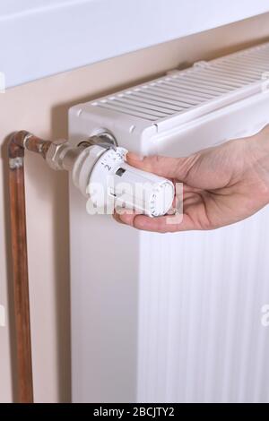 The woman's hand holds the radiator temperature control valve and sets the required value. White radiator and copper water pipes on the second backgro Stock Photo