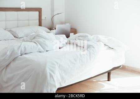 Working home quarantine self isolation lockdown concept. Laptop in unmade bed. Comfortable and cozy remote home office. Modern apartments with sunligh Stock Photo