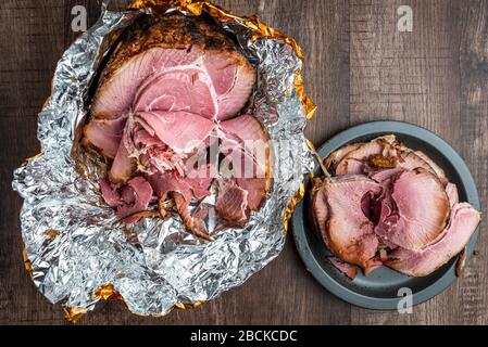 Spiral cut glazed and cooked ham in a foil wrapper on a wood table, black plate with slices Stock Photo