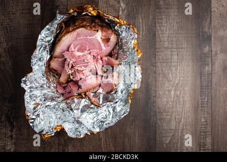 Spiral cut glazed and cooked ham in a foil wrapper on a wood table Stock Photo