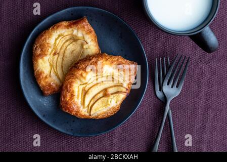 Fresh individual apple tarts on a black plate with forks, mug of milk, purple cloth background Stock Photo