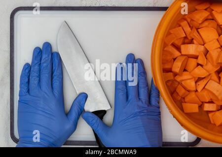 A man in a blue glove holds a knife, near cut and peeled sweet potatoes in a plastic bowl, safeguards and caution against the corona virus even in you Stock Photo