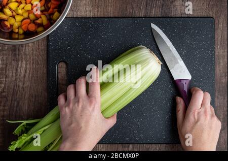 Woman’s hands, bunch of celery washed and ready to chop on a black cutting board with chef knife Stock Photo