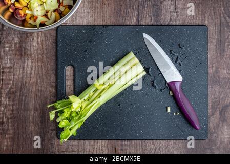 Celery stalks washed and ready to chop on a black cutting board, chef knife, wood table Stock Photo