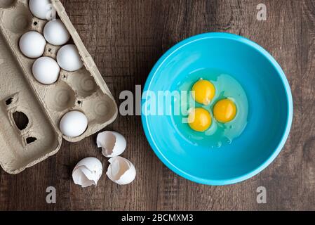 Three cracked eggs in blue mixing bowl, eggshells, whole eggs in cardboard carton, wood table Stock Photo