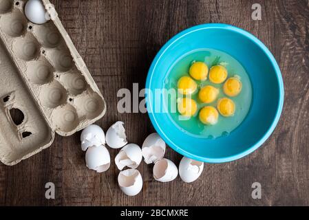 Eight cracked eggs in blue mixing bowl, eggshells, whole eggs in cardboard carton, wood table Stock Photo