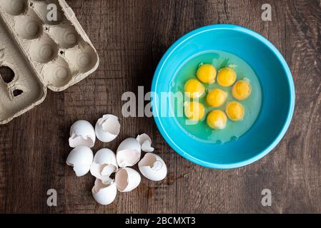 Eight cracked eggs in blue mixing bowl, eggshells, whole eggs in cardboard carton, wood table Stock Photo