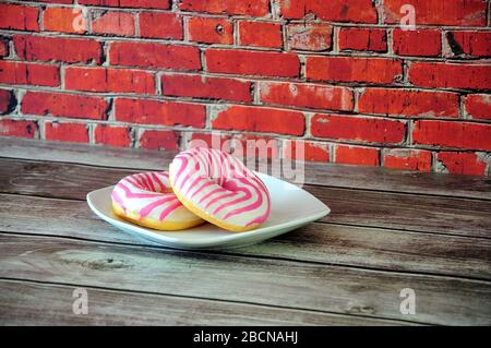 Two donuts in pink strawberry glaze on a white plate stand on a wooden table against a brick wall. Close-up. Stock Photo
