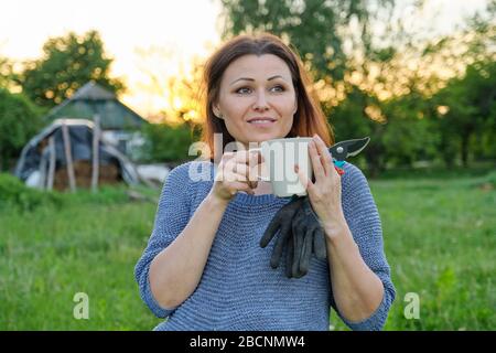 Beautiful mature woman gardener in gloves with secateurs with cup of herbal tea Stock Photo