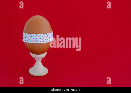 Egg standing on cup, on a soft red background. Easter egg tied with a festive ribbon. Stock Photo
