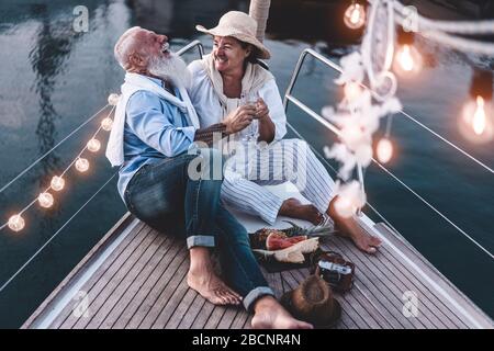 Senior couple cheering with champagne on a sailboat during summer vacation - Old people having fun together drinking and laughing - Joyful elderly lif