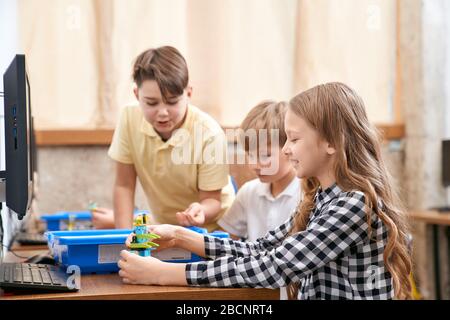 Interesting building kit for kids on table with computers. Side view of boys and girl creating toys. Science engineering. Nice interested friends chat Stock Photo