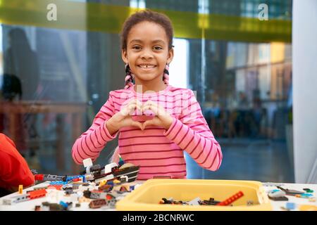 Front view of lovely smiling african girl standing, looking at camera and making heart with hands. Building kit for kids on table, children creating t Stock Photo