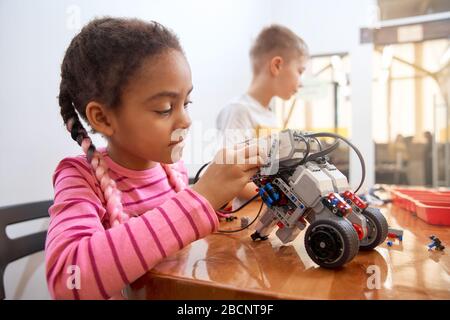 Building kit with colorful pieces for group of multiracial kids creating toys. Close up of concentrated african girl working on project, having positi Stock Photo
