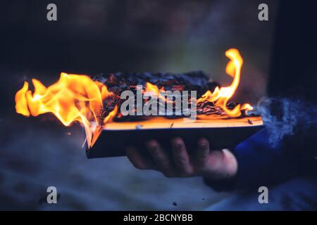 A man holds a book in his hand, burning with a bright strong flame and scattering light ashes through the air in the dark night. Stock Photo