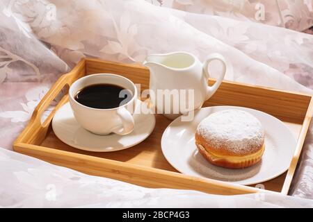 Morning breakfast in bed with fresh coffee, milk and doughnut in powdered sugar, in white dishes on a wooden tray. Horizontal view Stock Photo
