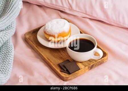 Morning breakfast in bed with fresh and hot coffee, a doughnut in powdered sugar and two slices of chocolate on a wooden tray against a backdrop of pi Stock Photo