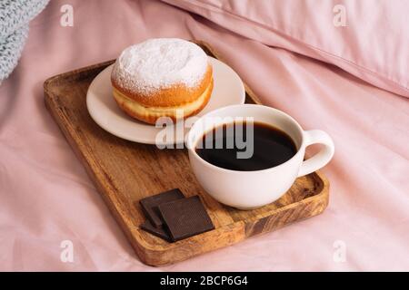 Morning breakfast in bed with fresh and hot coffee, a doughnut in powdered sugar and two slices of chocolate on a wooden tray against a backdrop of pi Stock Photo