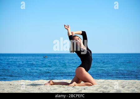 Beautiful woman practices yoga by the sea on a sunny day. The woman does stretching exercises. Stock Photo