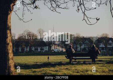 London, UK - March 22,2020: Rear view of two men sitting on the opposite sides of the bench watching people play ball in Broomfield Park, public park Stock Photo