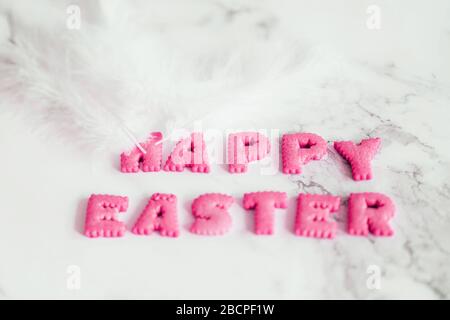Lettering from pink biscuits in form of letters and white fluffy plumelets on marble background. Happy Easter background, creative greeting. Stock Photo