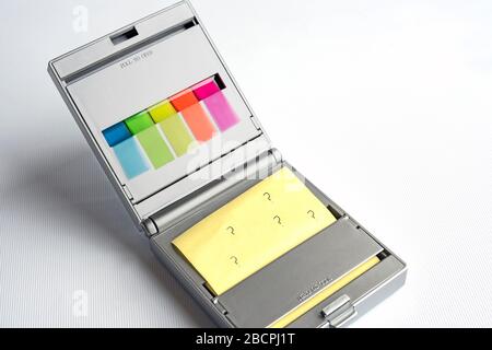 Silver dispenser of yellow postit notes and multi coloured sticky index highlighters against a white background, question marks written on postit note Stock Photo