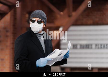 Horizontal shot of man uses virus preventive measures during coronavirus pandemic, wears face medical mask and rubber gloves, reads newspaper. Epidemi Stock Photo