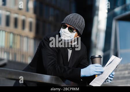 Young man at street with takeaway coffee and newspaper, wears protective medical mask and gloves, prevents spreading coronavirus, looks pensively asid Stock Photo