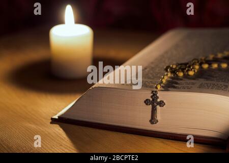 A candle, a bible and a cross a rosary on a wooden table Stock Photo