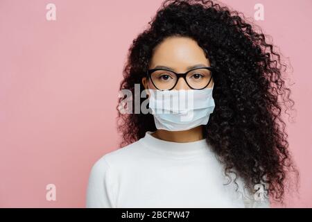 Covid-19, infectious virus. Close up shot of young woman with curly bushy hair, wears transparent glasses and medical disposable mask, cares about her Stock Photo