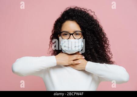 Sick curly haired woman touches neck, suffers from suffocation and shortage of breathing, wears medical mask to avoid virus infection, isolated on pin Stock Photo