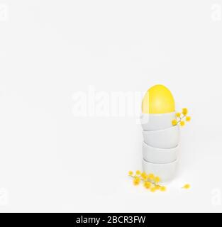 Easter composition with a yellow egg in a stand and acacia flowers on a white background. Minimal Easter concept. Copy space. Side view. Stock Photo