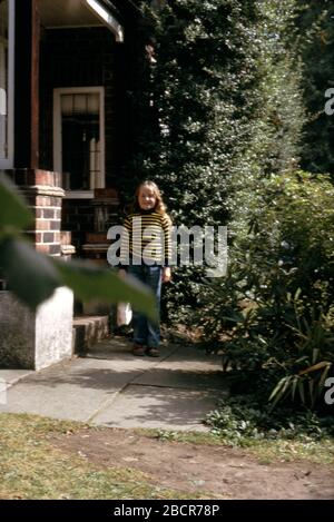 A young girl wearing a yellow and blue striped Breton sweater and blue patched jeans, carrying a Mickey Mouse bag with presents in, in the front garden of her family home, UK 1974 Stock Photo