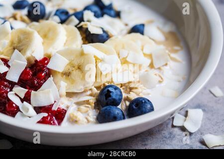 Bircher muesli with berries, banana, jam and coconut in a white bowl, gray background. Stock Photo
