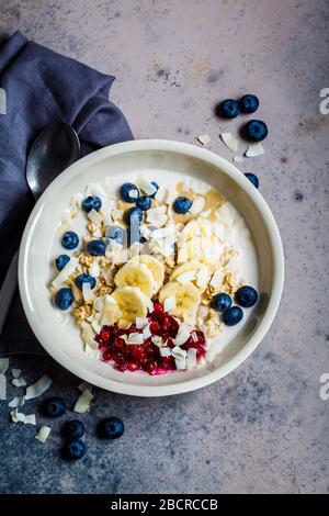 Bircher muesli with berries, banana, jam and coconut in a white bowl, gray background. Stock Photo