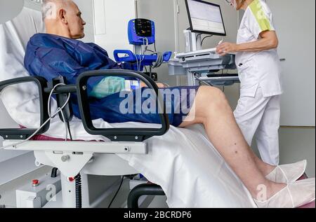 Curious attentive senior male on modern medical chair inside modern hospital in France resting after infection virus coronavirus optimistic view and nurse preparing data in medical computer network Stock Photo