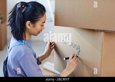 Side view portrait of smiling Asian woman writing on cardboard box labeling for moving out to new house, copy space Stock Photo