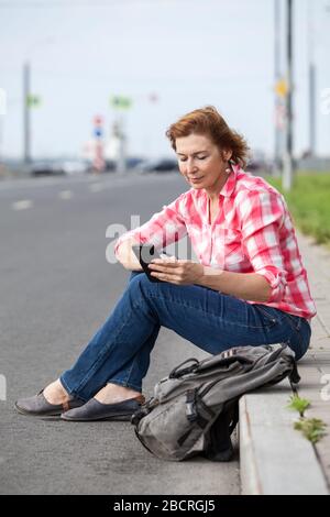 Mature Caucasian woman using smartphone for call for a taxi, sitting on a roadside with backpack Stock Photo
