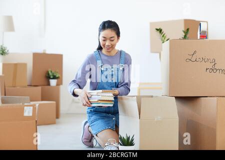 Full length portrait of young Asian woman packing books to cardboard boxes and smiling happily excited for moving to new house or dorm, copy space Stock Photo