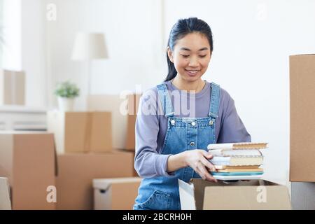 Portrait of young Asian woman packing books to cardboard boxes and smiling happily excited for moving to new house or dorm, copy space Stock Photo