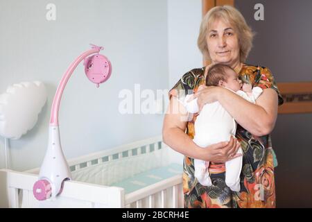 Portrait of grandma carrying a newborn infant in her arms, stands near white crib in bedroom, looking at camera Stock Photo