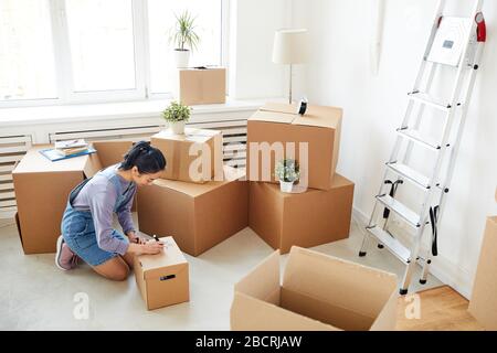 Wide angle background of young Asian woman packing cardboard boxes in empty white room, moving, relocation and house decor concept, copy space Stock Photo