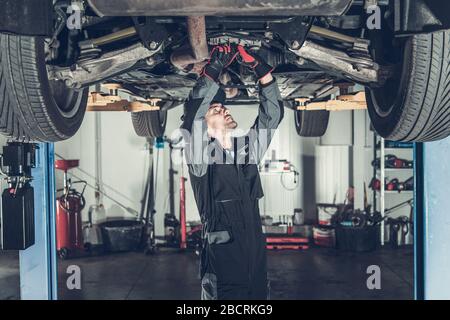 Caucasian Car Mechanic Under Vehicle Looking For Potential Issues with a Drivetrain. Automotive Industry. Stock Photo