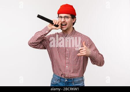 Funny and passionate cool hipster guy in red beanie and glasses singing rap song acting excited, holding microphone, having fun at karaoke with Stock Photo