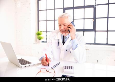 Shot of senior male doctor using mobile phone and talking with his patient while sitting behind his laptop in the doctor’s office. Stock Photo